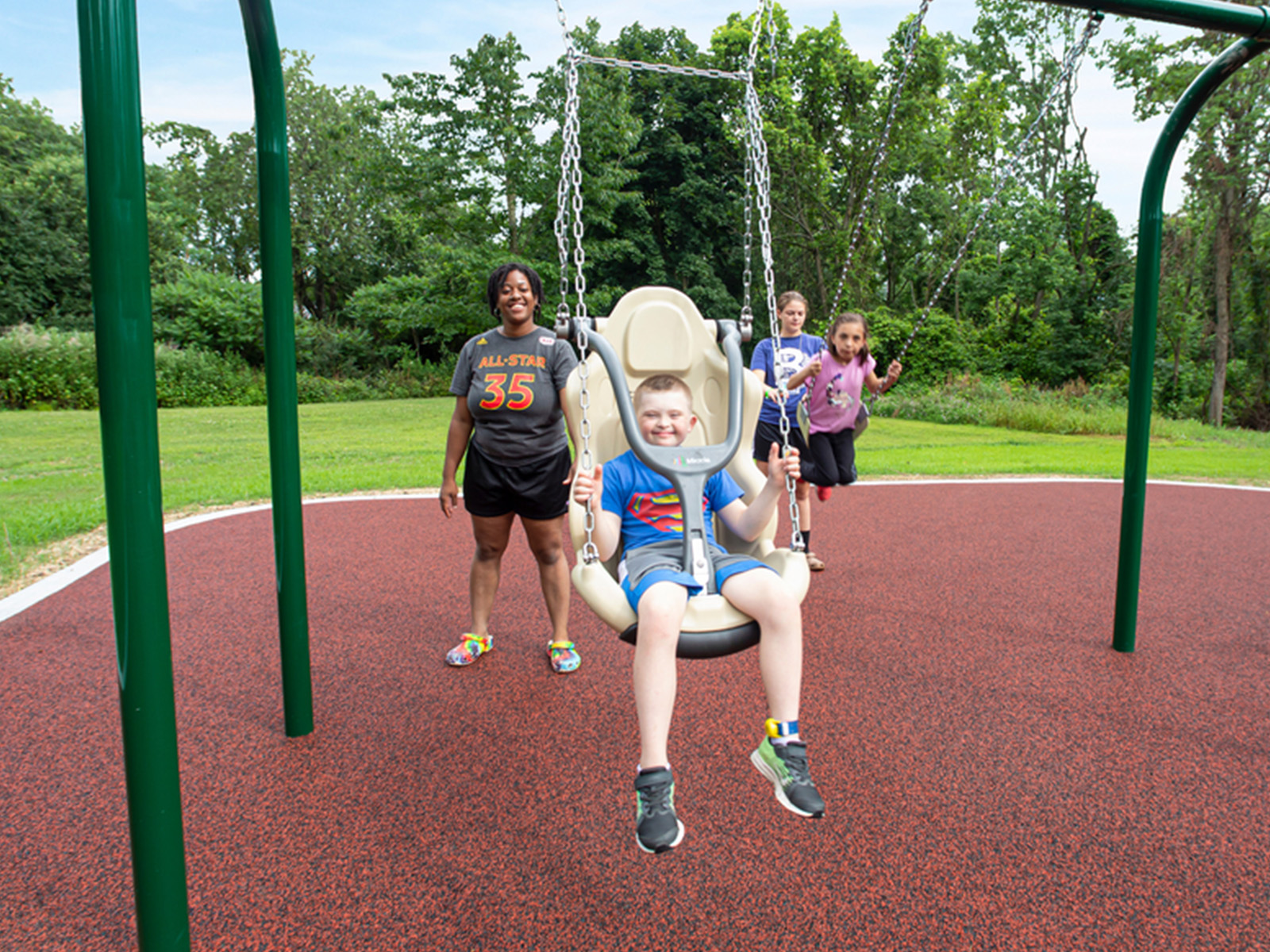 Smiling boy in an adaptive swing seat, with an adult and girls on swings behind him.