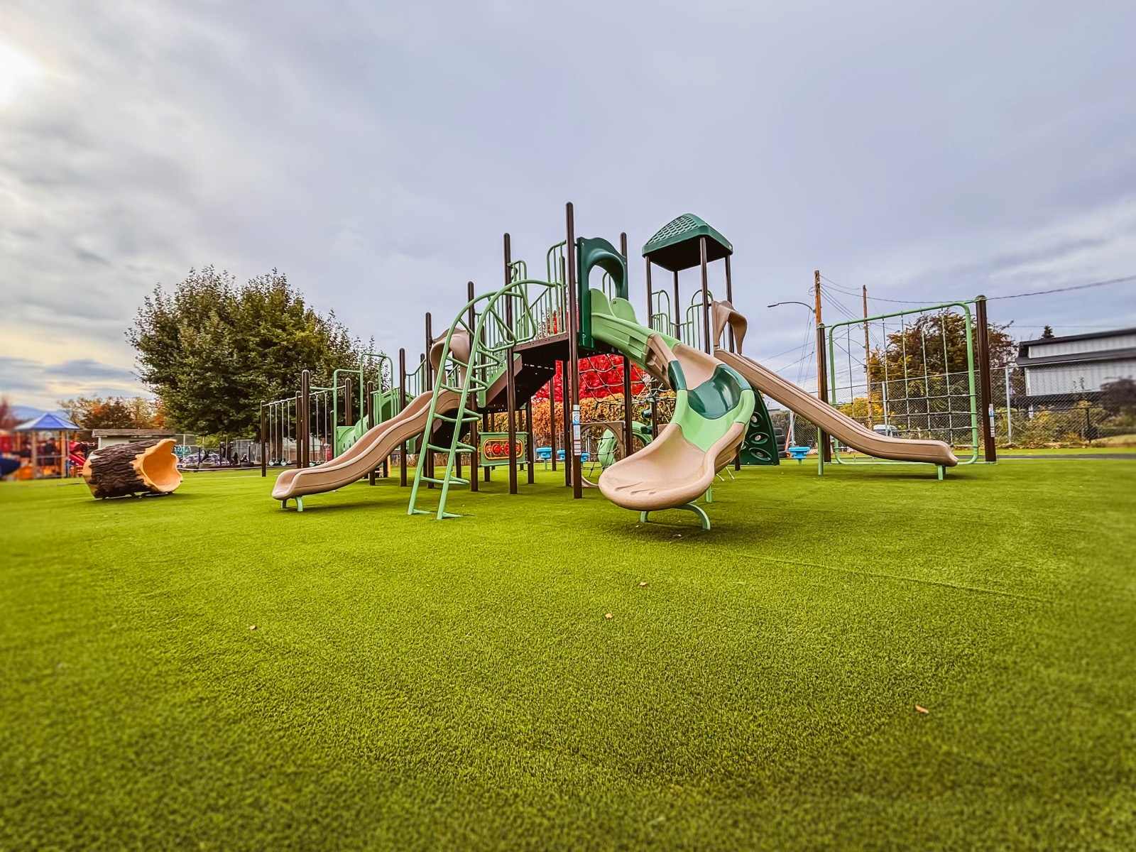 Brown and green play structure with tan slides and a hollow log on artificial turf under cloudy skies.